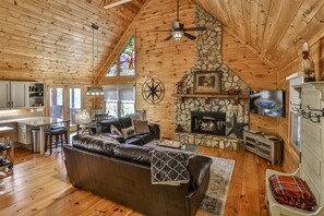 A cozy cabin interior with a stone fireplace, leather sofa, wooden ceiling, and a television.
