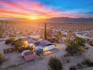 A desert landscape with a small cluster of buildings, including a white house with solar panels, surrounded by sparse vegetation and a clear sky.