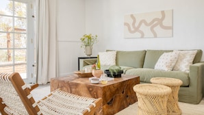 Un salon avec un canapé vert, une table basse en bois, des fauteuils en osier et une vue sur l’extérieur, visible à travers une porte vitrée.