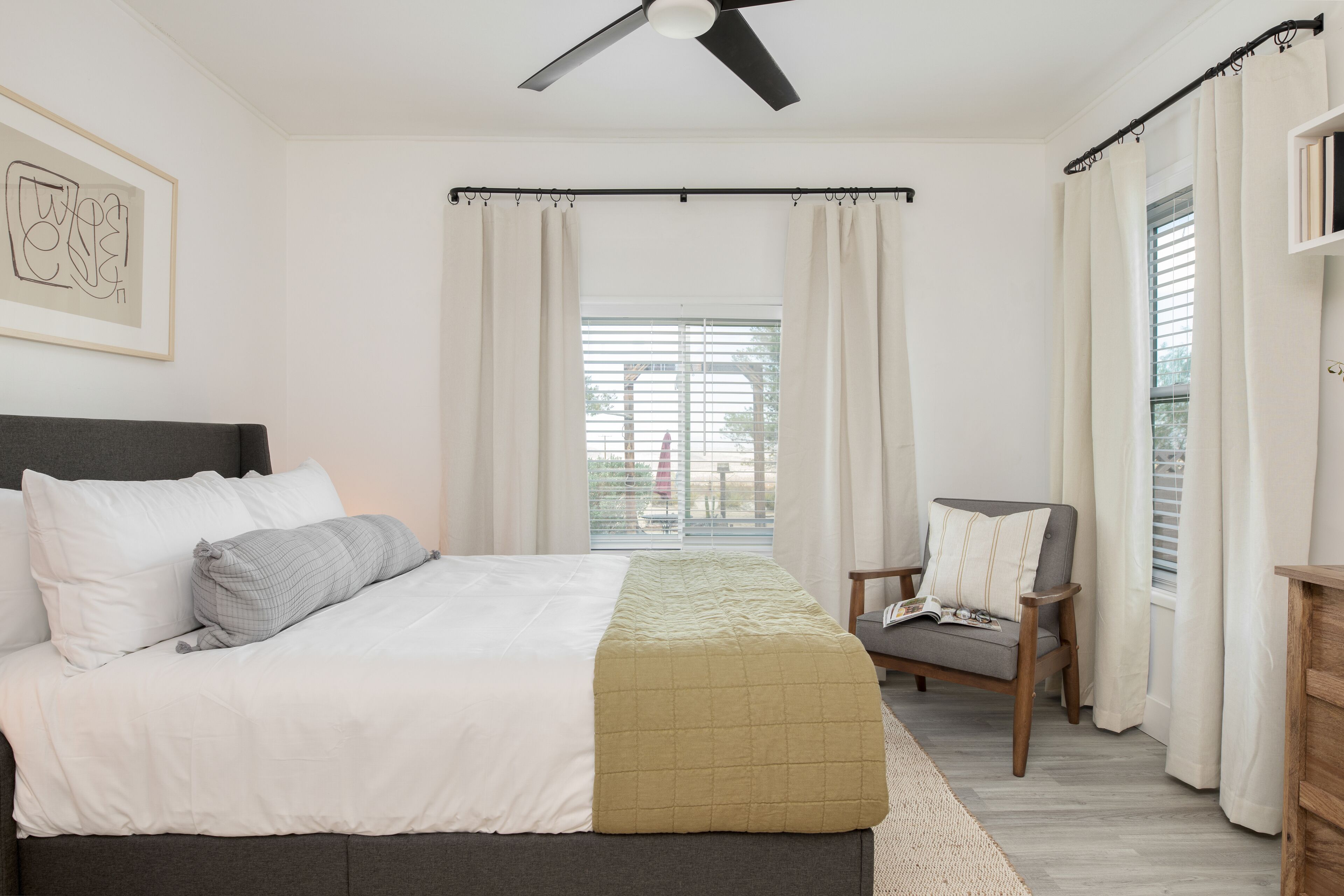 Un dormitorio con cama, una silla, ventilador de techo y ventana con cortinas.