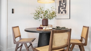 Un coin repas avec une table ronde, deux chaises en bois, un vase avec des feuilles vertes, un bol de raisins, une assiette de fromage et une œuvre d’art encadrée accrochée au mur.