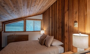 Um quarto com teto inclinado, paredes de painel de madeira, uma cama com almofadas, uma mesa de cabeceira, um abajur e uma janela com persianas.