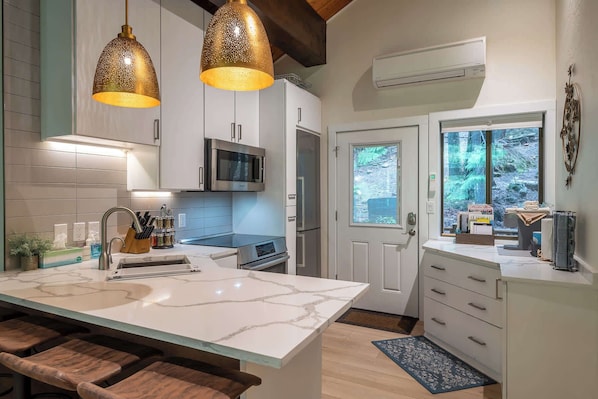 A modern kitchen with a marble island, stainless steel appliances, pendant lighting, and a view through a window.