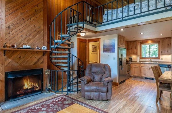 A wooden interior with a fireplace, a spiral staircase, a leather chair, a dining table, and a kitchen area.