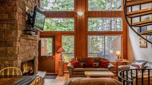A cozy living room with a stone fireplace, leather furniture, large windows, and a wooden staircase. - Escape to Ptarmigan Village 90 (Whitefish)