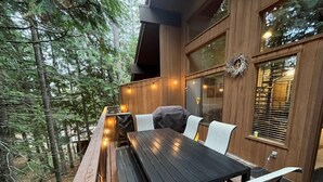 An outdoor wooden deck with a table and chairs, surrounded by trees. - Escape to Ptarmigan Village 90 (Whitefish)