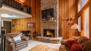 A cozy living room with a fireplace, wooden walls, a television, and a balcony. - Escape to Ptarmigan Village 90 (Whitefish)