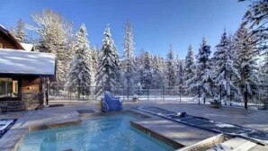 A swimming pool with a stone deck, surrounded by snow-covered trees and a wooden building. - Escape to Ptarmigan Village 90 (Whitefish)