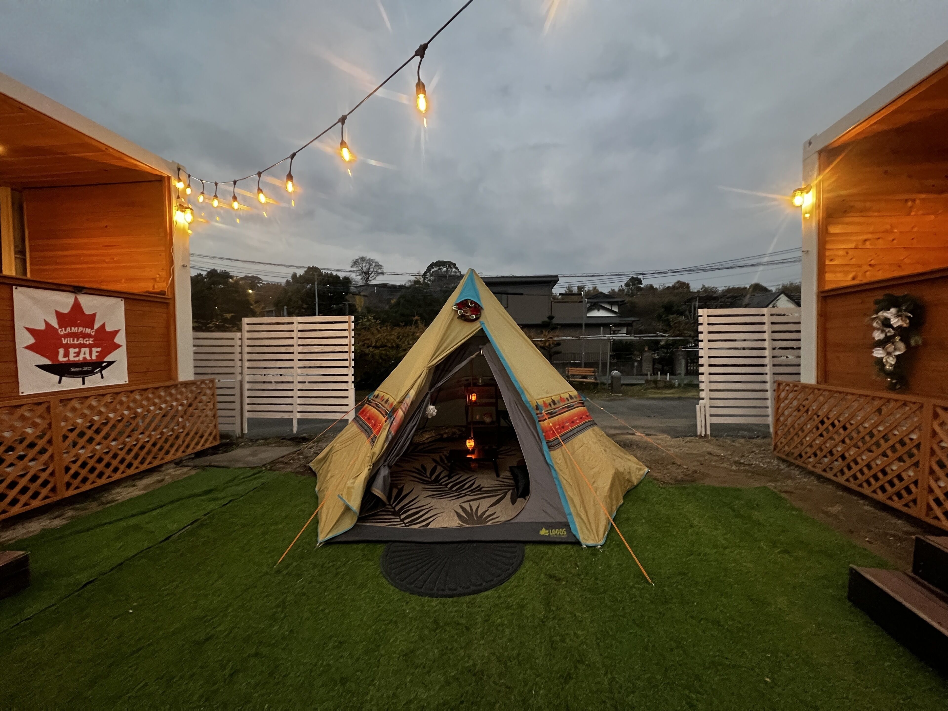 A tent set up in an outdoor area with artificial lighting, surrounded by wooden structures and a sign indicating a camping village.