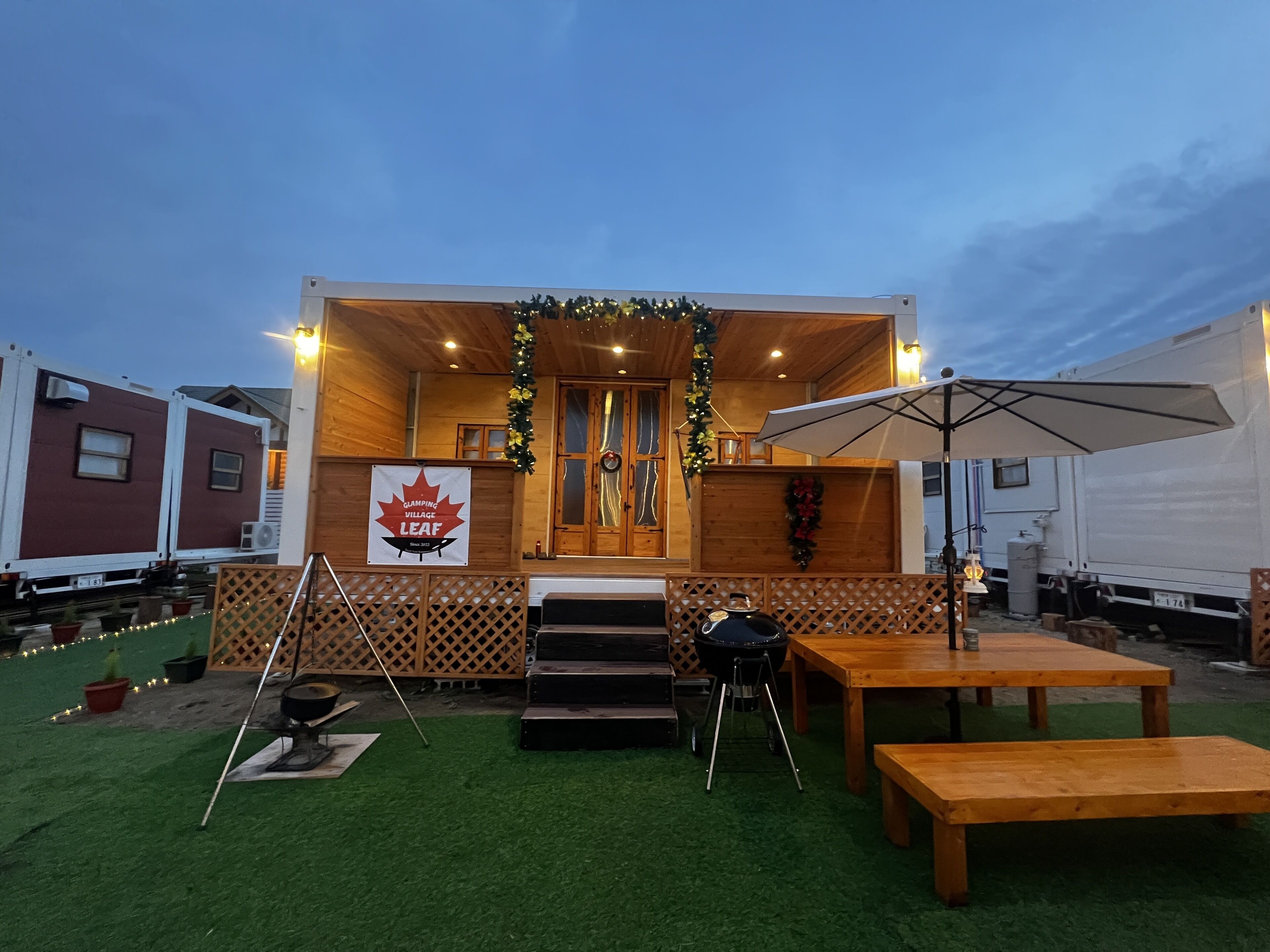 A wooden outdoor dining area with a table and benches, a grill, and a sign for Glamping Village Leaf.