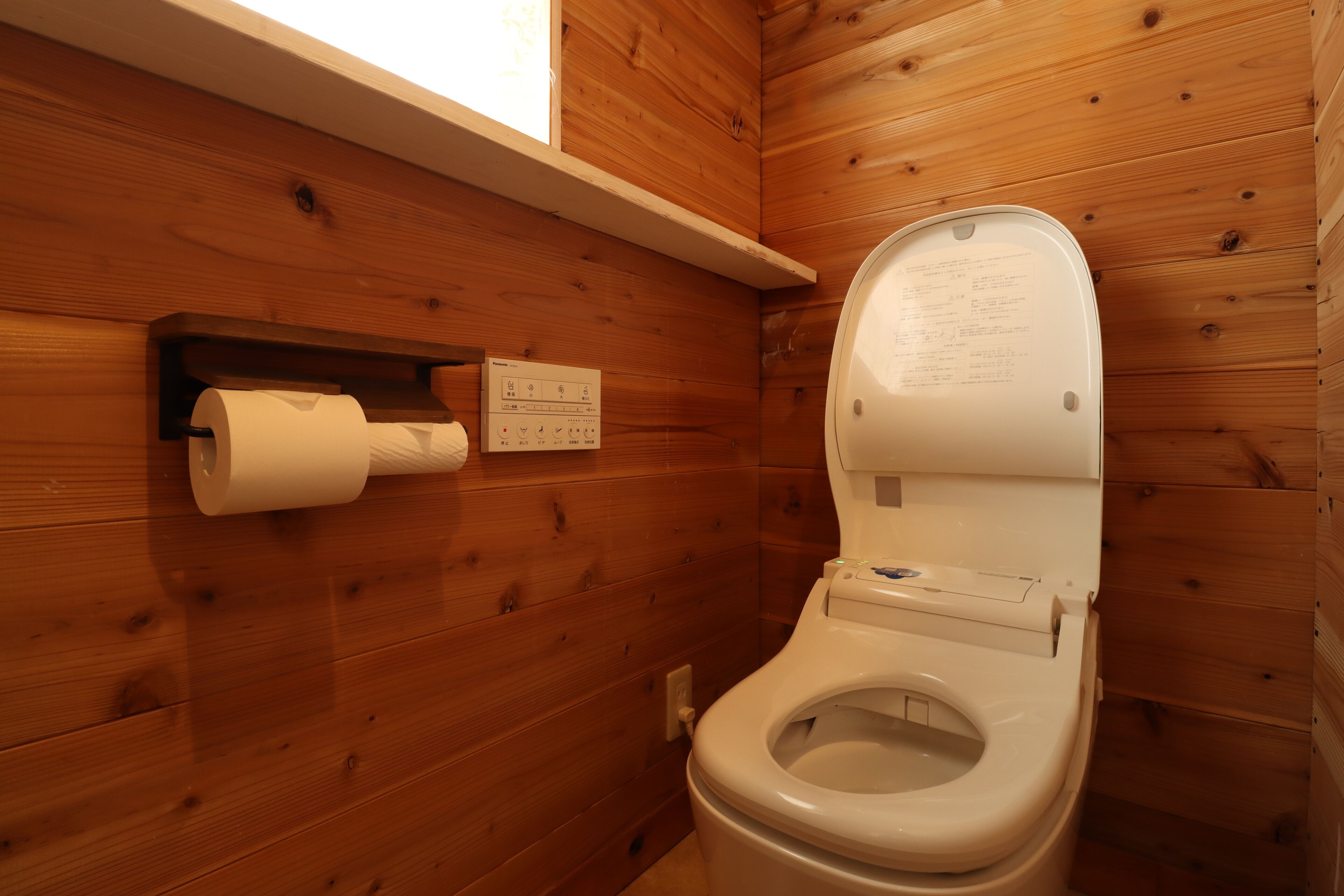 A wooden-paneled bathroom with a toilet, a paper towel dispenser, and a window above the toilet.
