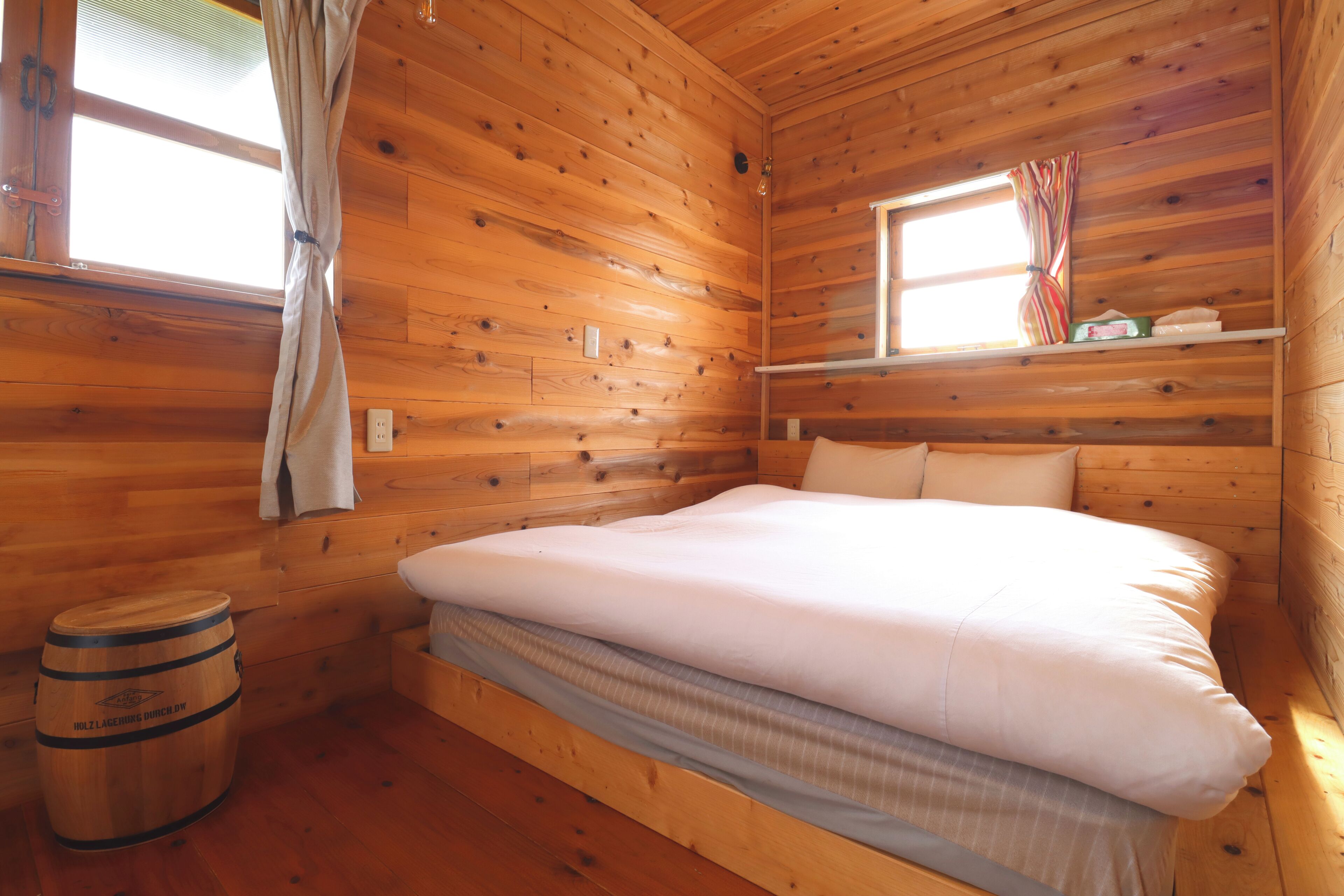 Una habitación de cabaña de madera con una cama, una ventana con cortinas, una repisa con objetos y un barril.
