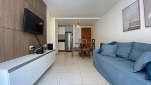 A modern living room with a flat-screen TV, a blue sofa, a dining area with a table and chairs, and a kitchen in the background. - Bela Hospedagem - Unique Beach Bessa (João Pessoa)