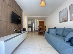 En moderne stue med et fladskærms-tv, en blå sofa, et spiseområde med et bord og stole, og et køkken i baggrunden.