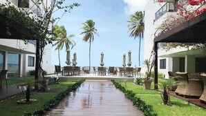 A hotel courtyard with a wooden walkway, outdoor seating, palm trees, and a view of the ocean.