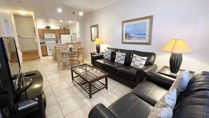 Ein Wohnzimmer mit schwarzem Ledersofa, einem Glastisch und einem Essbereich mit Holzstühlen und einem kleinen Tisch.