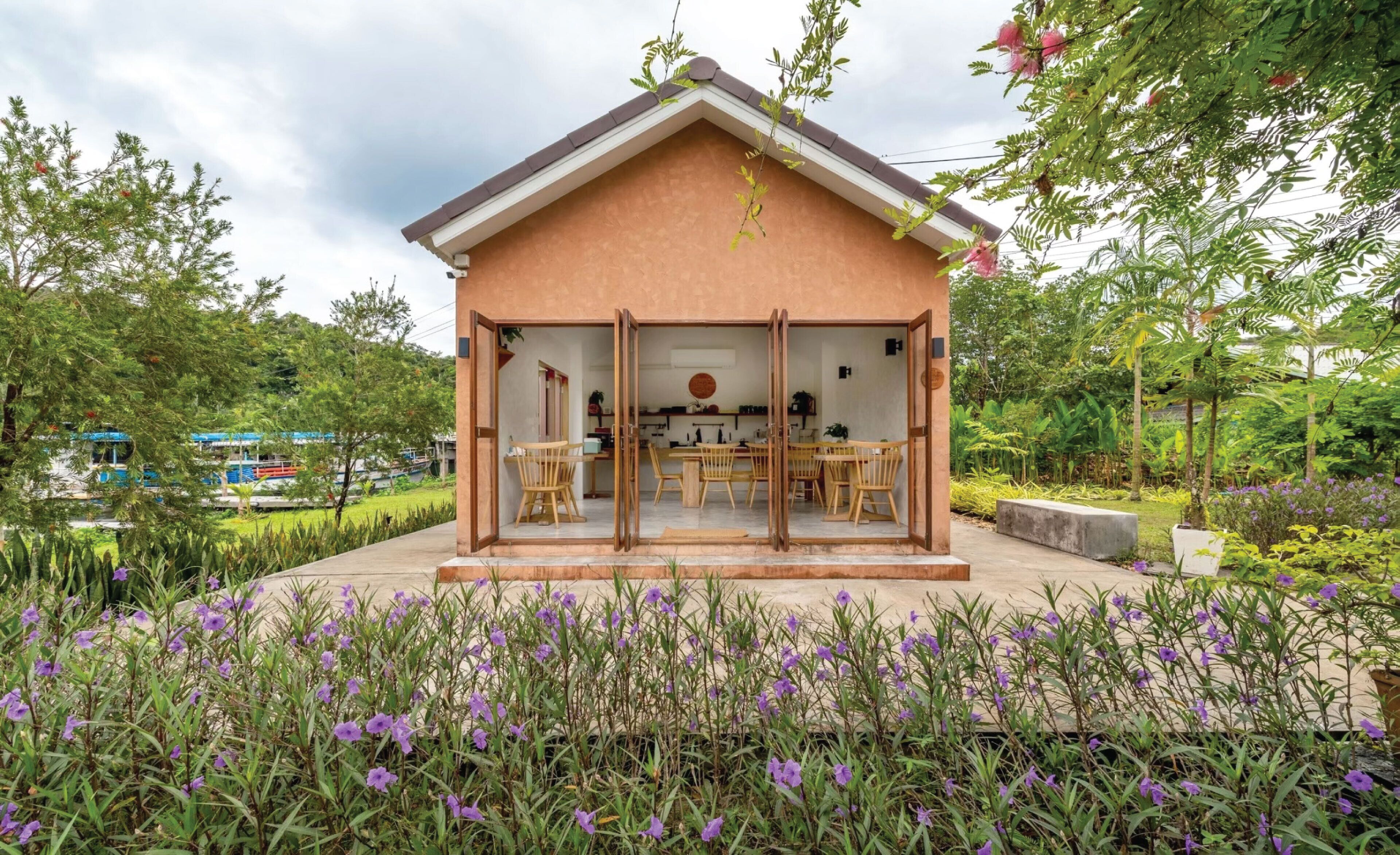 Une petite maison à aire ouverte, comprenant un coin repas et une cuisine, entourée d’un jardin où poussent des fleurs violettes.
