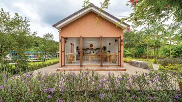 Une petite maison à aire ouverte, comprenant un coin repas et une cuisine, entourée d’un jardin où poussent des fleurs violettes.