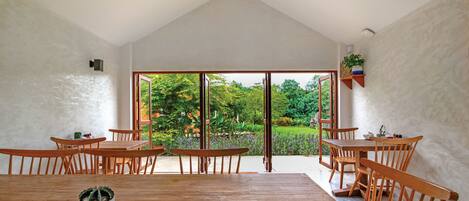 A dining area with wooden tables and chairs, a ceiling fan, and a view of a garden through large glass doors.
