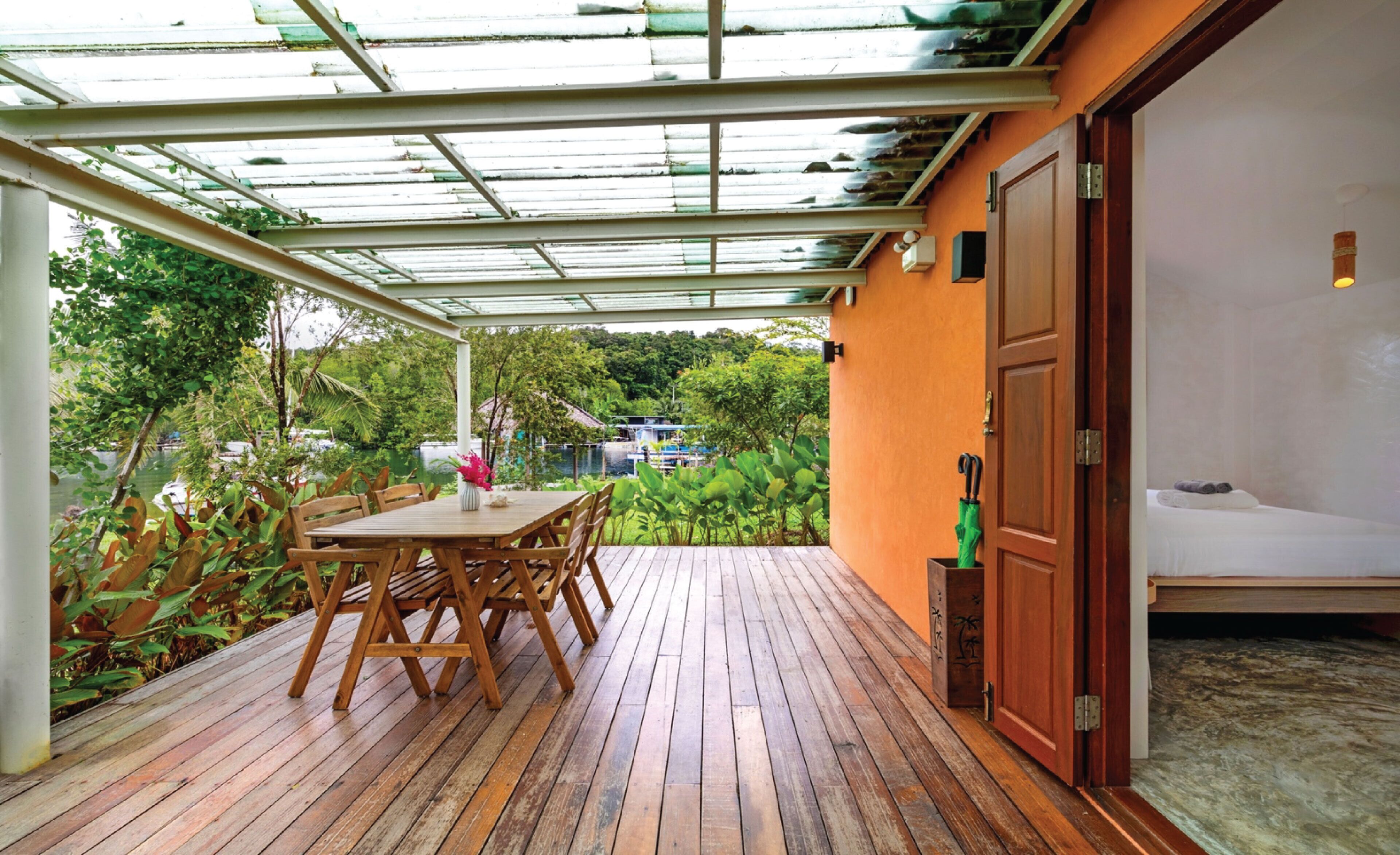 A wooden deck with a table and chairs, a glass ceiling, and a view of greenery and a building in the background.