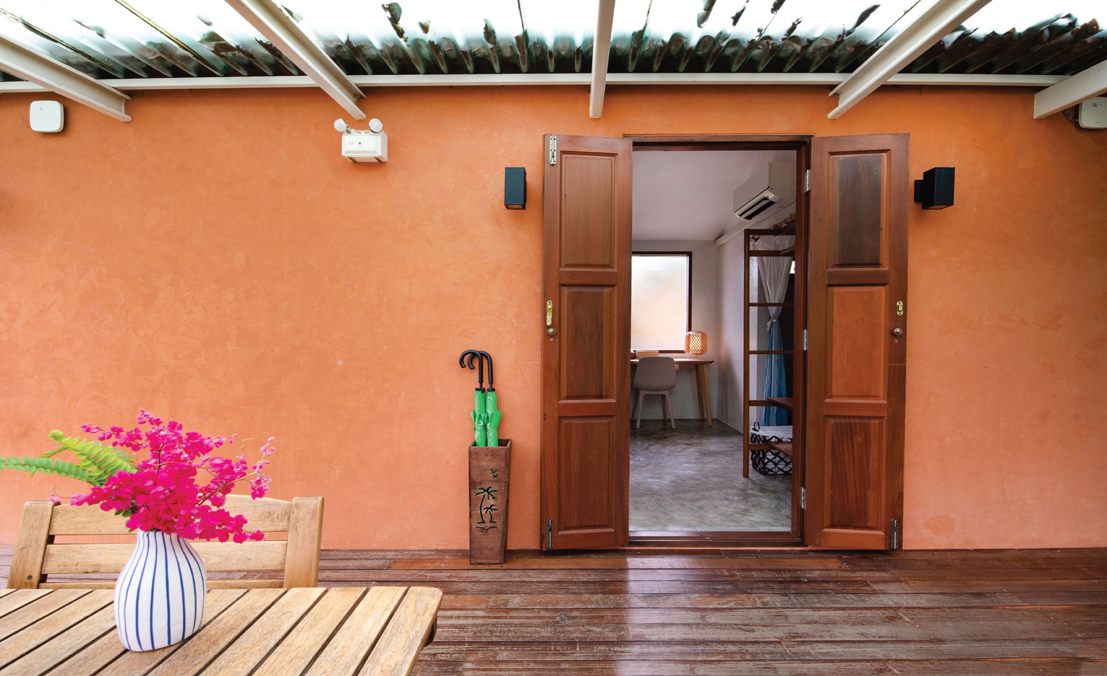 A wooden bench with a striped vase of pink flowers, a wooden door, and a wooden bench with a woven basket.