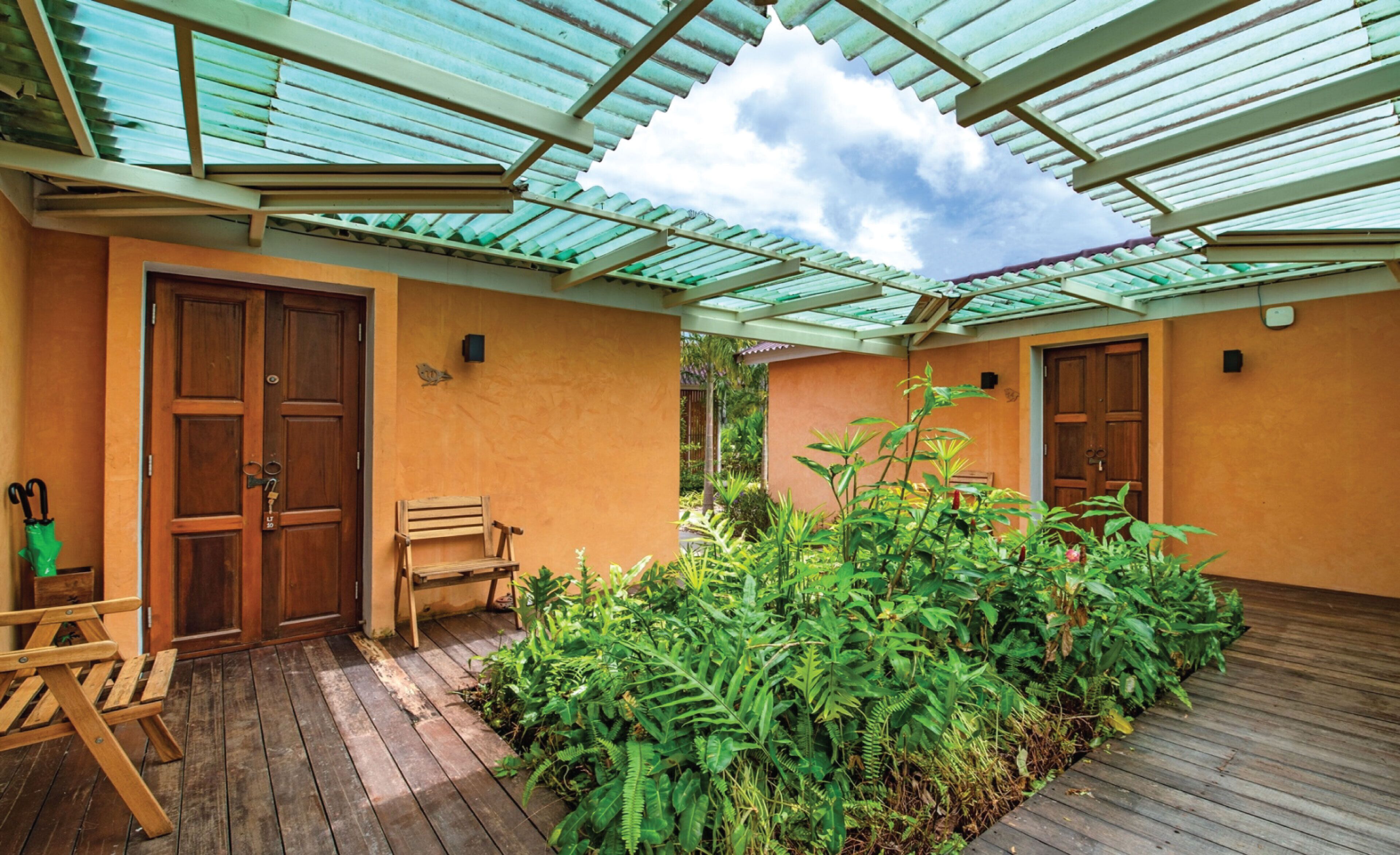 A covered outdoor area with wooden flooring, a bench, and lush greenery.