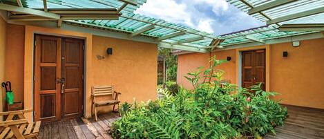 A covered outdoor area with wooden flooring, a bench, and lush greenery.