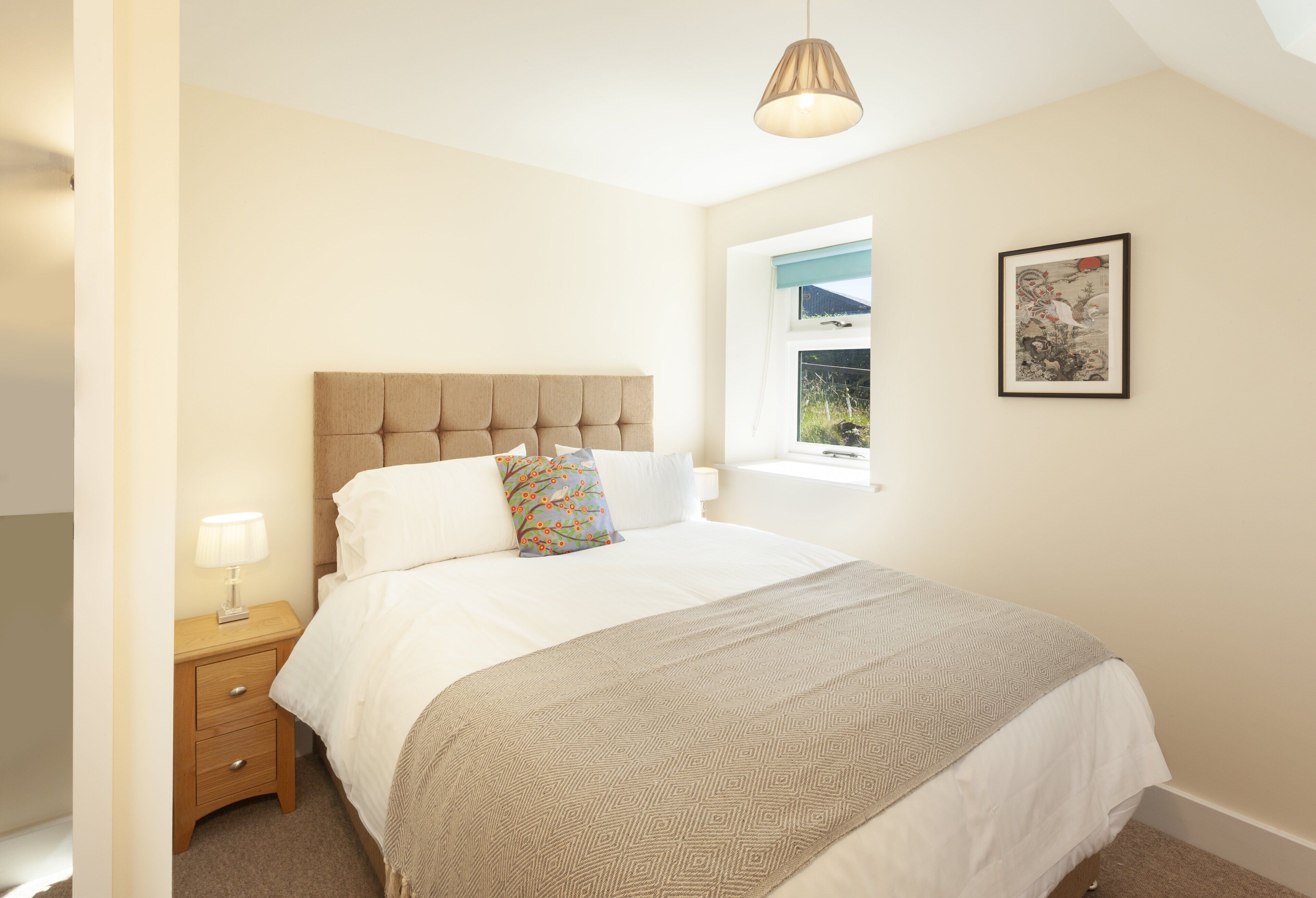 A bedroom with a bed, a bedside table, a lamp, a window with a view, and a framed picture on the wall.