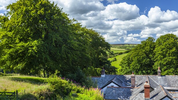 A rural landscape with a house featuring a sloped roof and chimneys, surrounded by lush greenery and trees. - Dashel Cottage, Countisbury (Lynmouth)