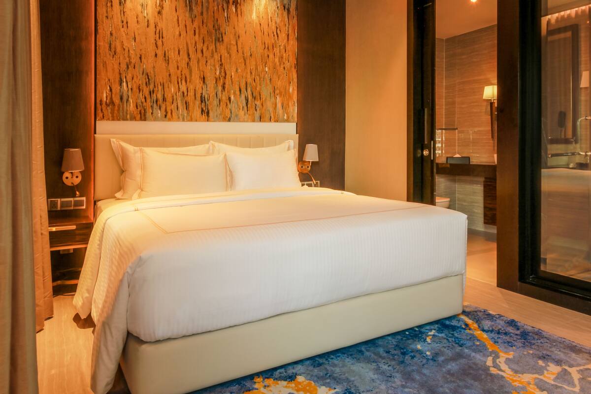A large bed with white linens, a wooden textured wall behind it, and a glass door leading to a bathroom.
