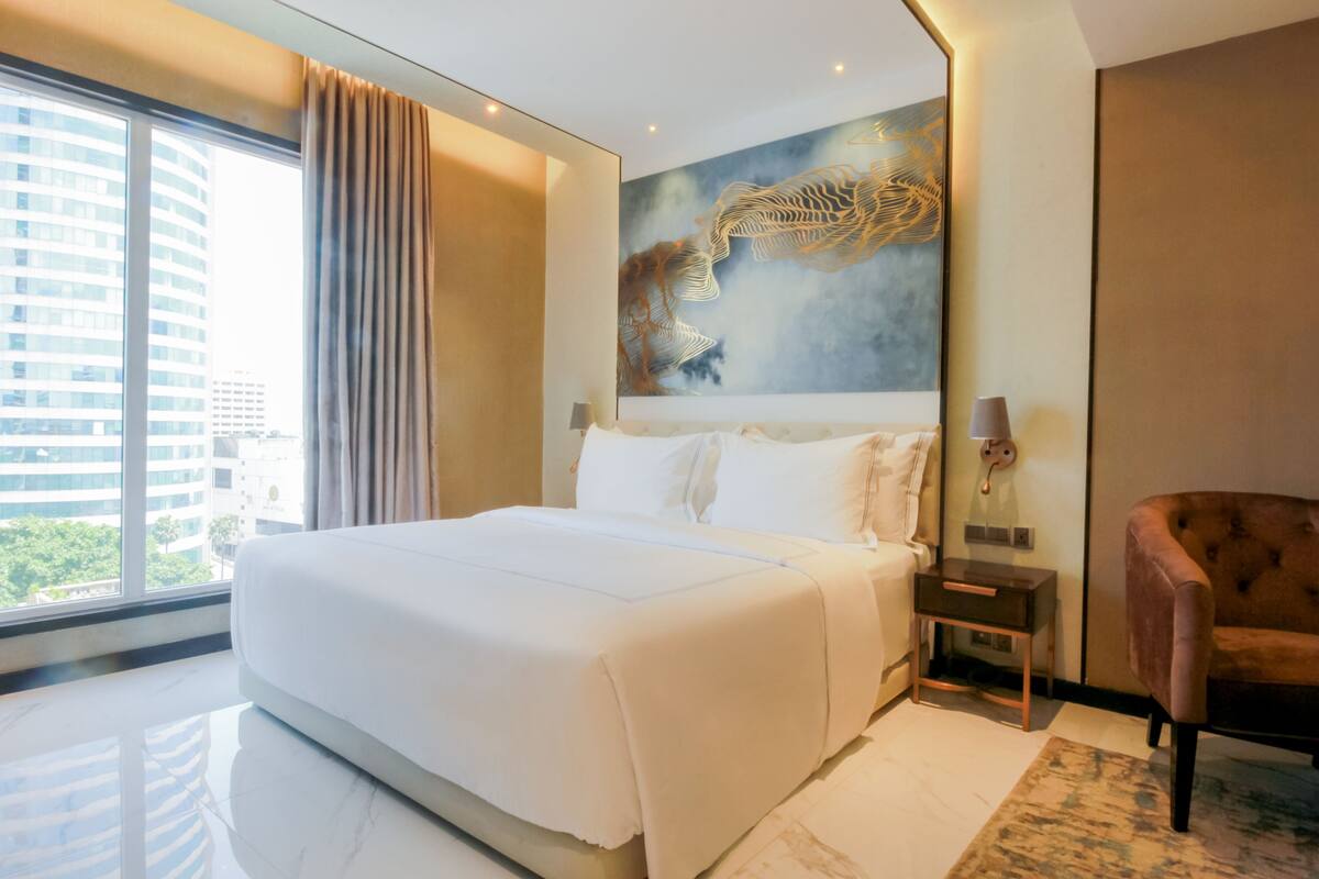 A modern hotel room with a large bed, a nightstand, a chair, and a large window offering a city view.