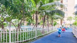 Un espace de jeux pour enfants doté d’une surface bleue en caoutchouc, entouré d’une clôture blanche, de palmiers et d’un bâtiment visible au loin.