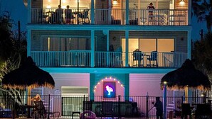 Una casa frente al mar de dos pisos con balcones, un letrero de neón y sombrillas de paja.