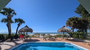 Zona de piscina con vista a la playa, palmeras y sombrillas de paja.