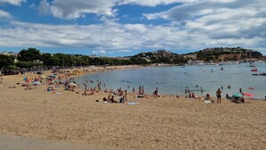 Ein Strand mit sonnenbadenden und schwimmenden Personen, einem klaren Himmel und Gebäuden im Hintergrund.