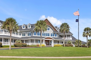 Uma casa grande de dois andares, com varanda, várias janelas e um mastro com a bandeira americana.
