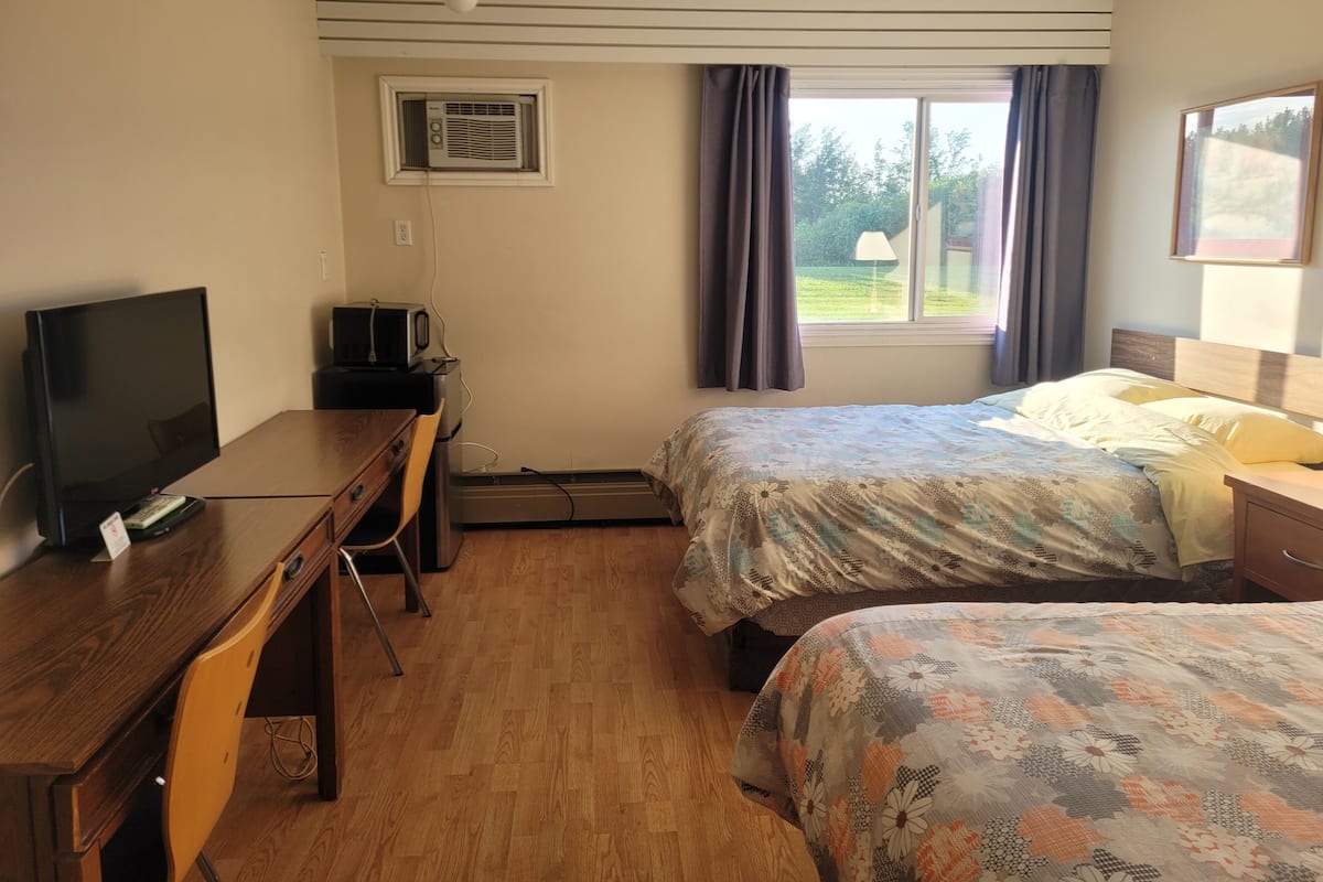 A hotel room with a wooden desk, a television, a bed with floral bedding, a window with curtains, and an air conditioning unit.