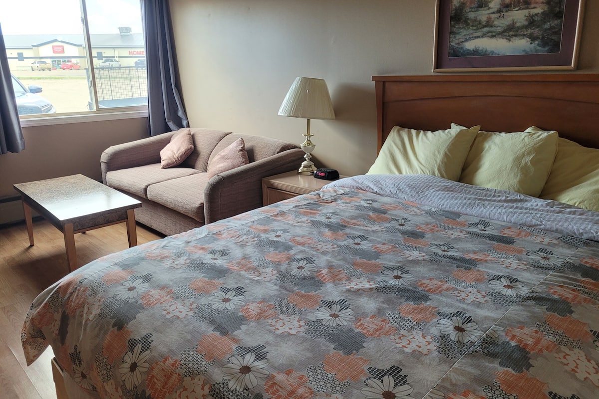 A hotel room with a bed, a chair, a small table, a lamp, and a painting on the wall.