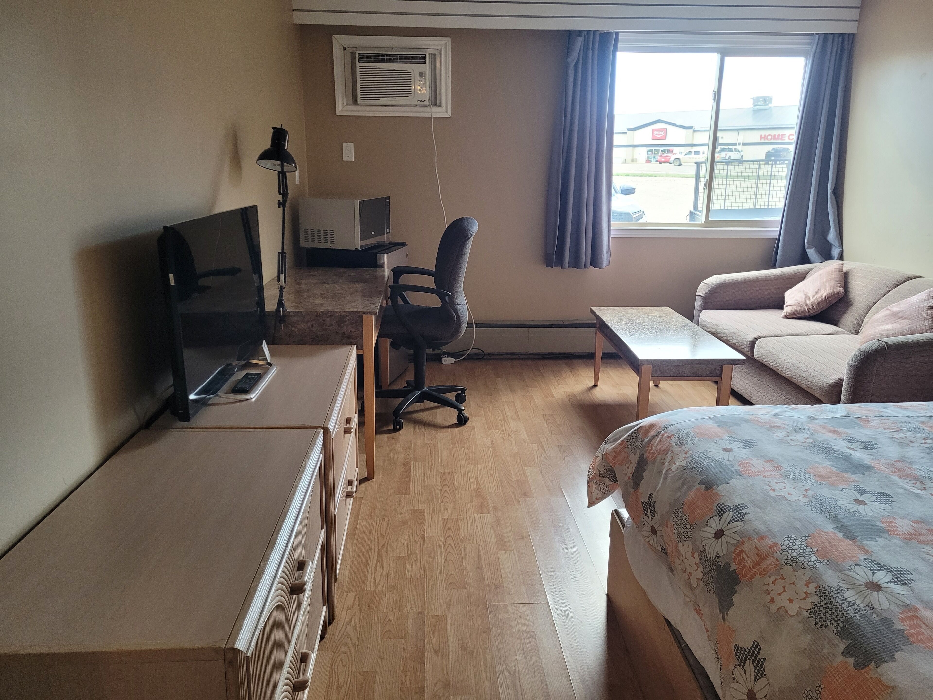 A hotel room with a bed, a desk with a computer, a chair, a sofa, and a window with curtains.