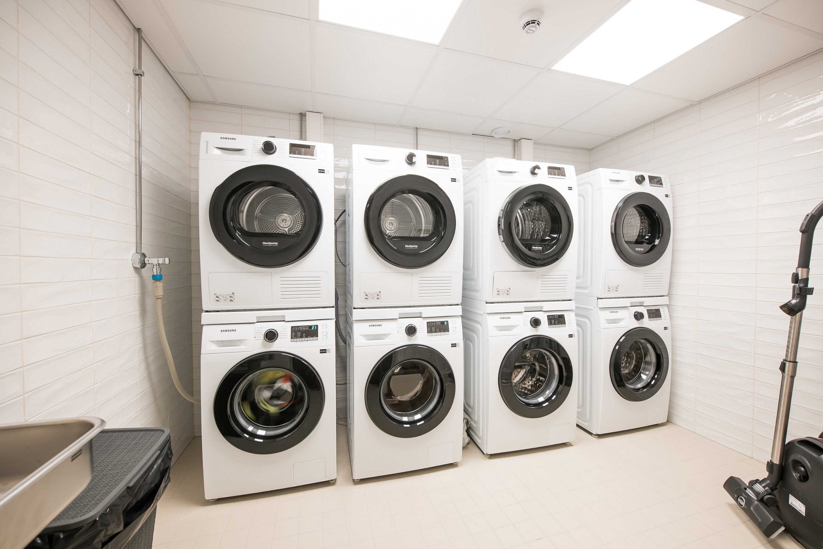 A laundry room with six stacked front-loading washing machines, a metal sink, and a vacuum cleaner.