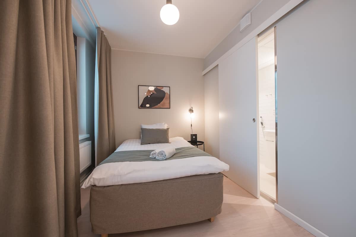 Un dormitorio con cama, mesita de noche, lámpara de pared, puerta y ventana con cortinas.