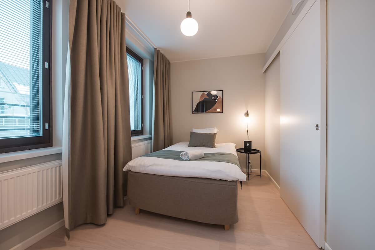 Un dormitorio con cama, mesita de noche, lámpara, ventana con cortinas y un cuadro colgado en la pared.