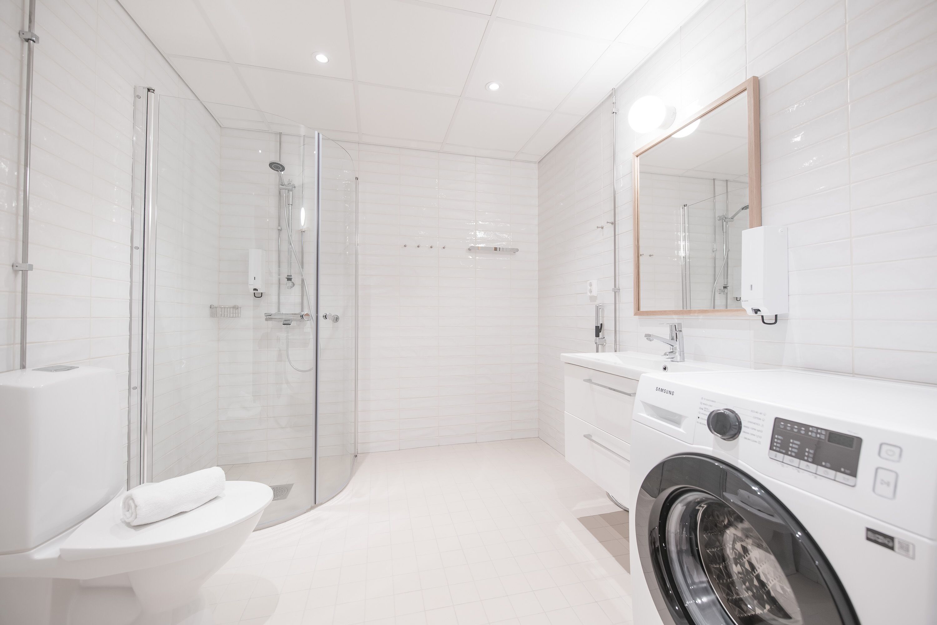 A modern bathroom with a glass-enclosed shower, a toilet, a washing machine, and a large mirror.