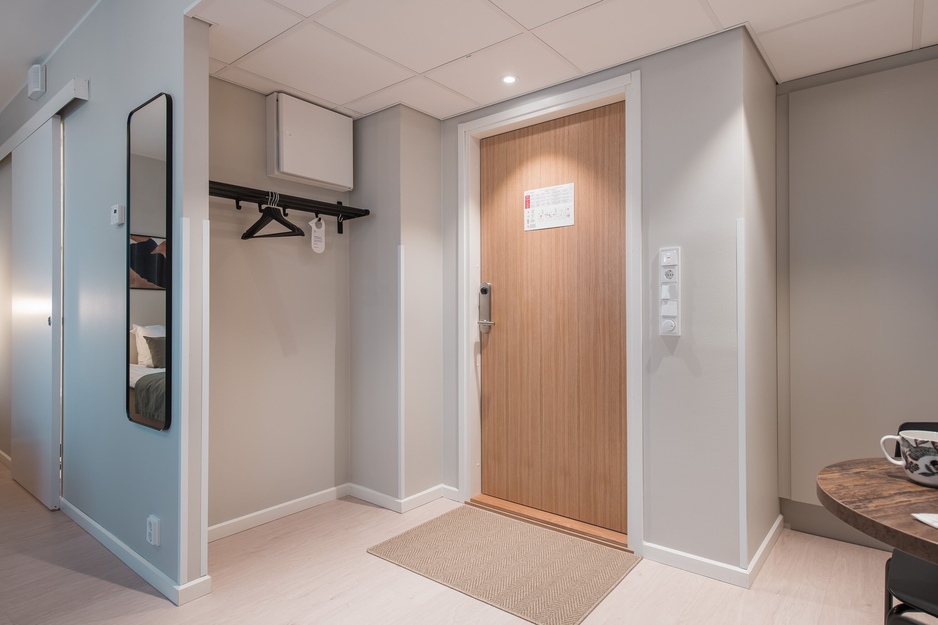 A hotel room with a wooden door, a mirror, a coat rack, and a small table with a cup.