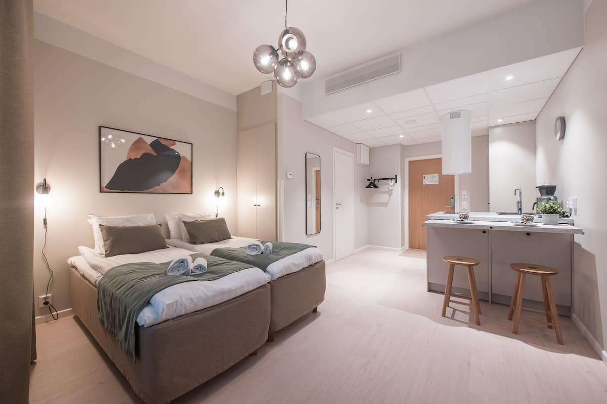 Una habitación de hotel moderna con una cama, lámparas de noche, un escritorio con taburetes y una zona de cocina.
