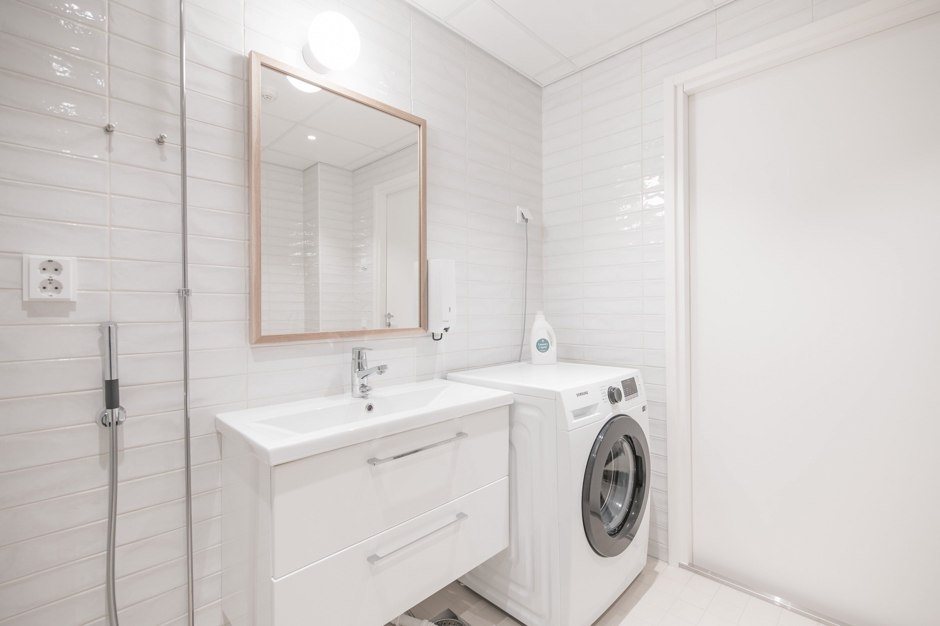 A modern bathroom with white tiles, a sink, a mirror, and a washing machine.