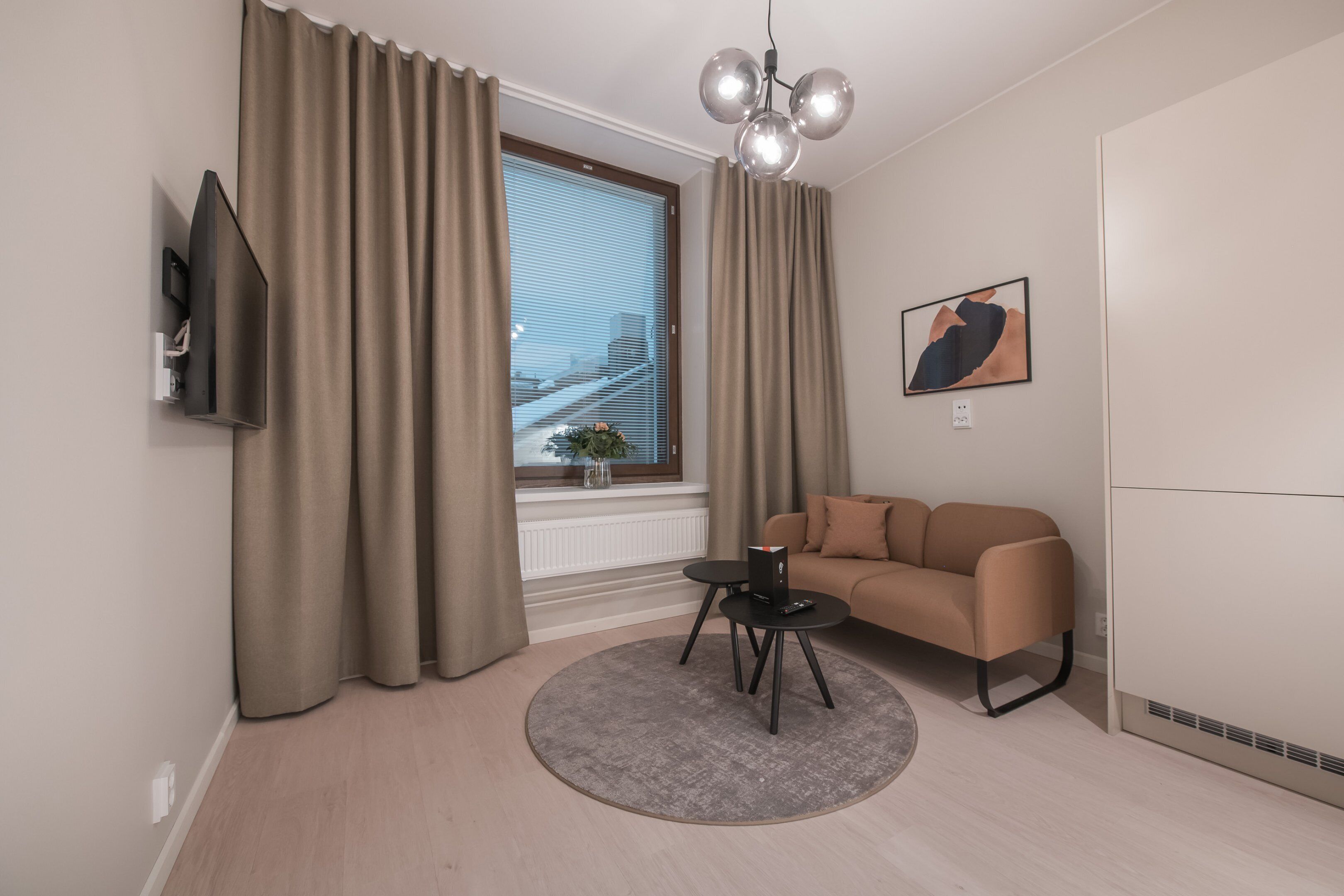 Una sala de estar moderna con un sofá, una mesita redonda, un televisor de pantalla plana montado en la pared y una ventana con cortinas.