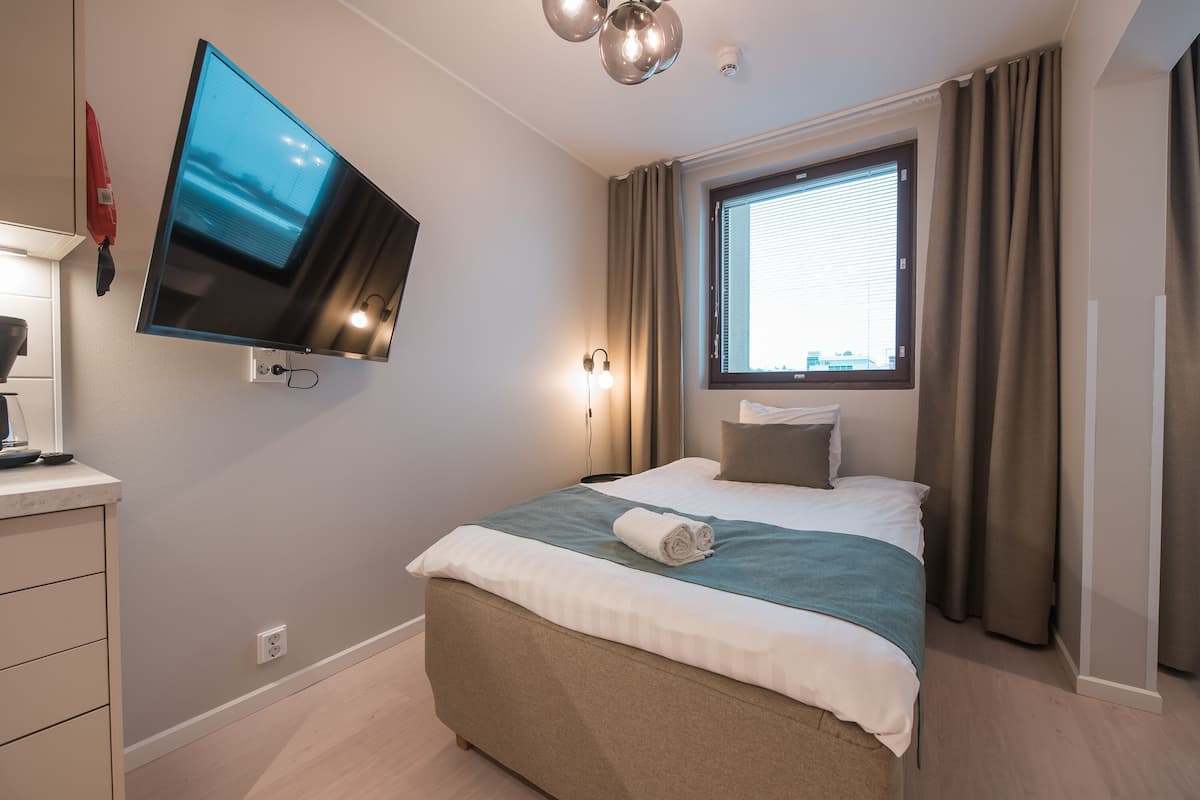 Una habitación de hotel moderna con una cama grande, un televisor de pantalla plana en la pared, una ventana con cortinas y una mesita de noche con lámpara.