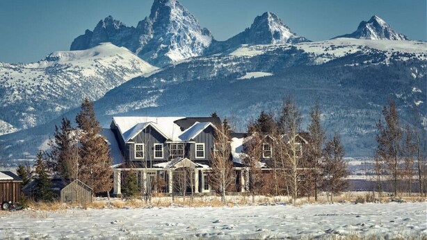 Una casa con techo nevado, rodeada de árboles y montañas al fondo.