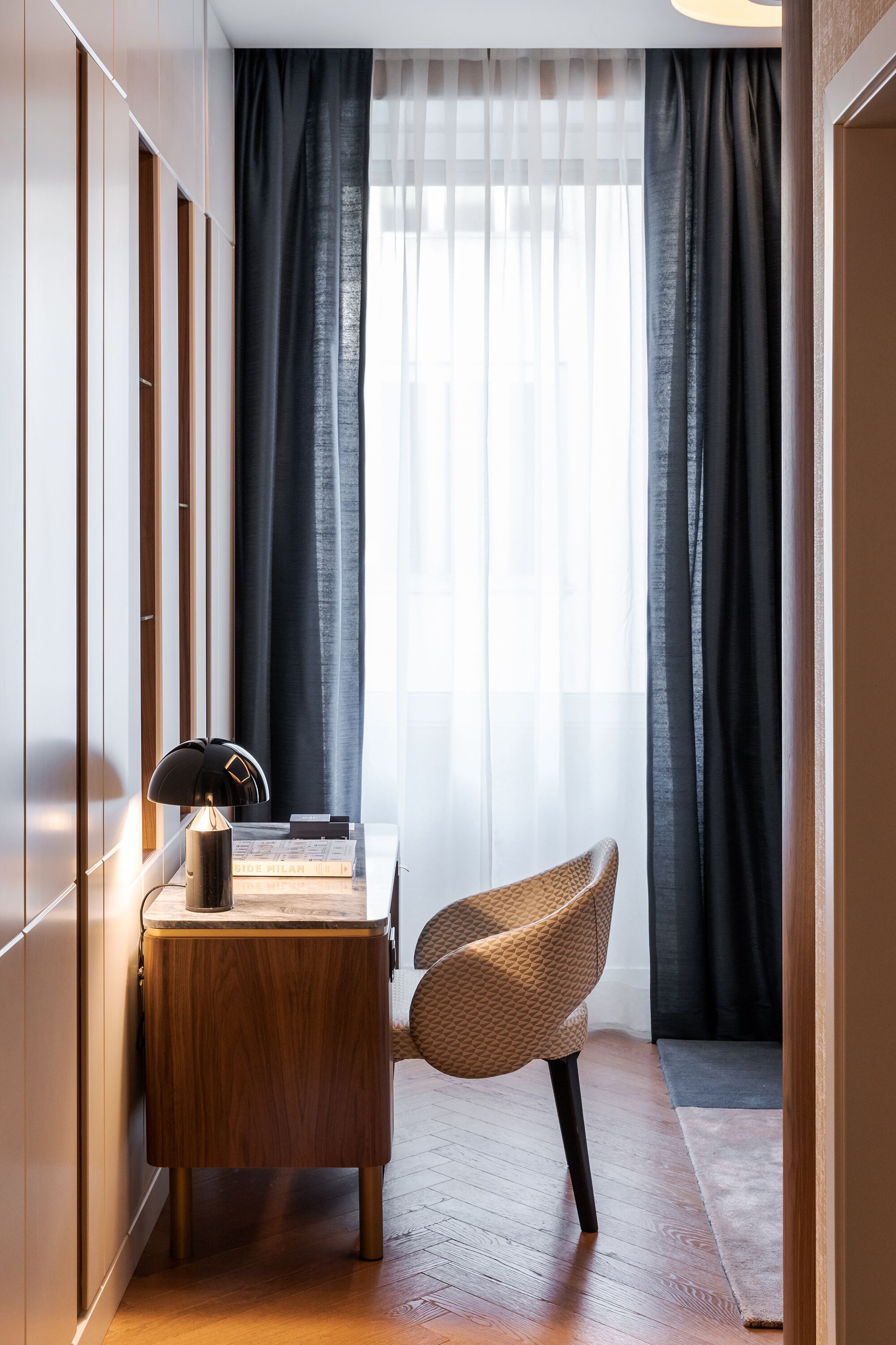 Una habitación de esquina con un escritorio de madera, una silla y un ventanal con cortinas traslúcidas.